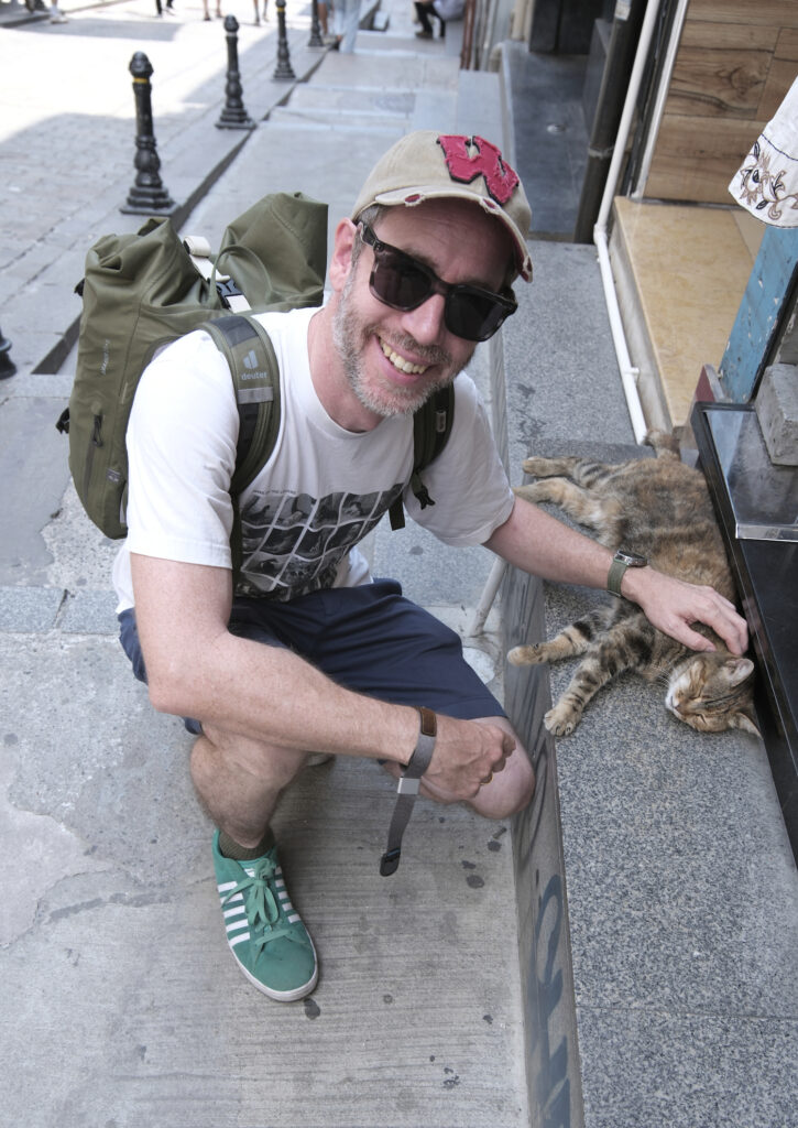 Picture of the photographer in shorts, sunglasses and cap while petting a street cat chilling on the sidewalk.